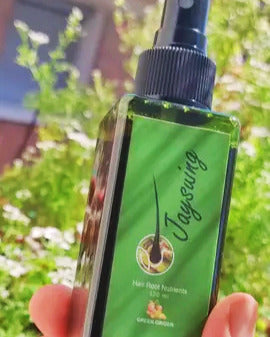 Green Ginger - Spray Estimulante Capilar botella de producto sostenido por una mano con flores de fondo en un dia soleado de españa