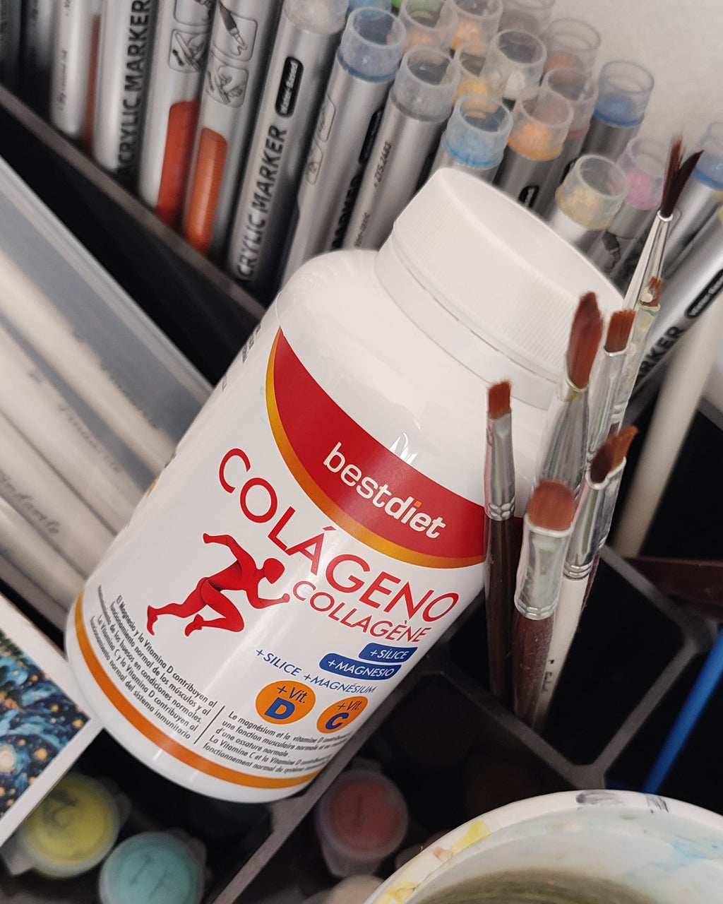 frasco de colageno encima de un organizador de pinceles, pinturas y marcadores de colores